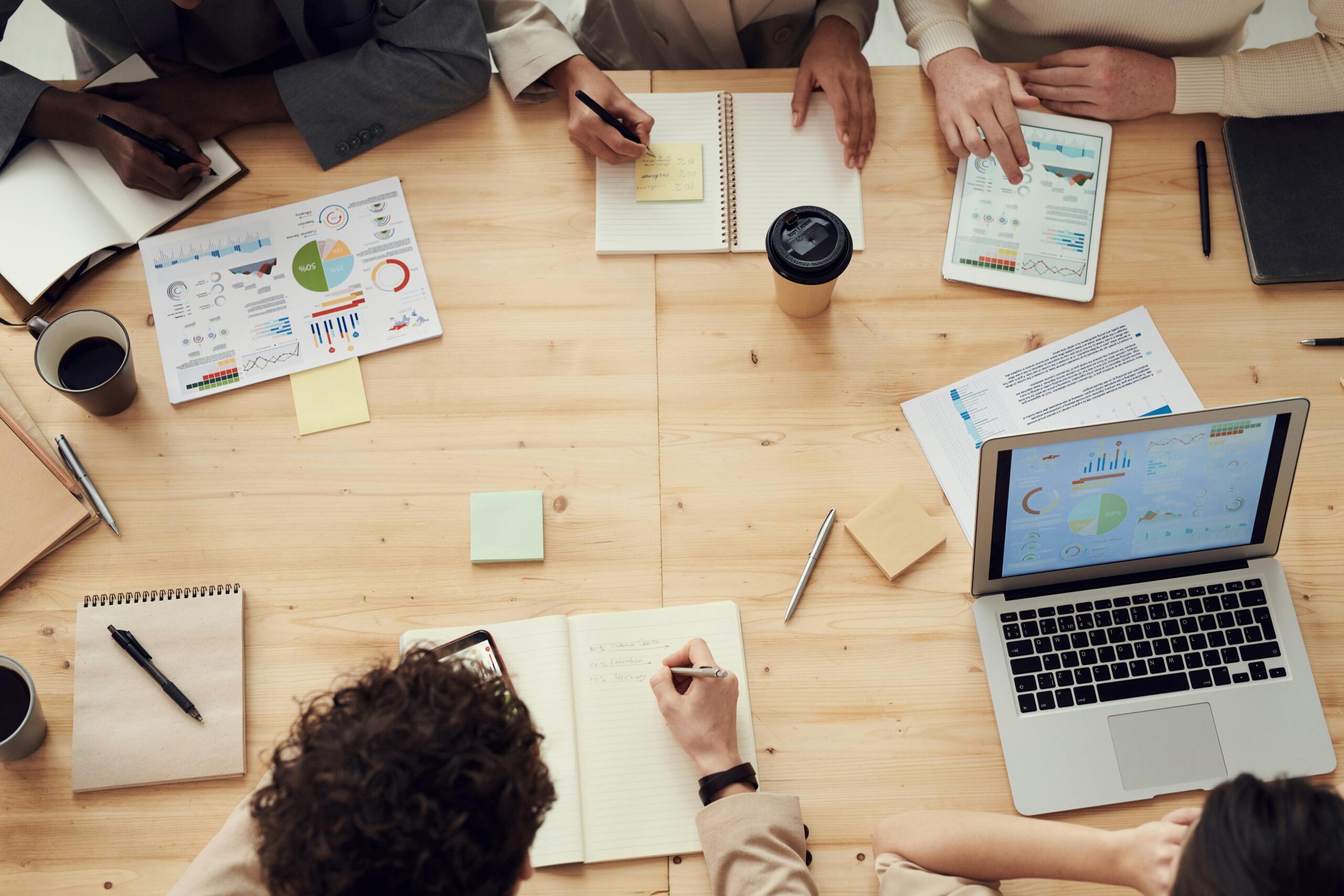 A group of people sitting at a wooden table with laptops, tablets, notebooks, and printed charts, collaborating on business strategy and analytics.