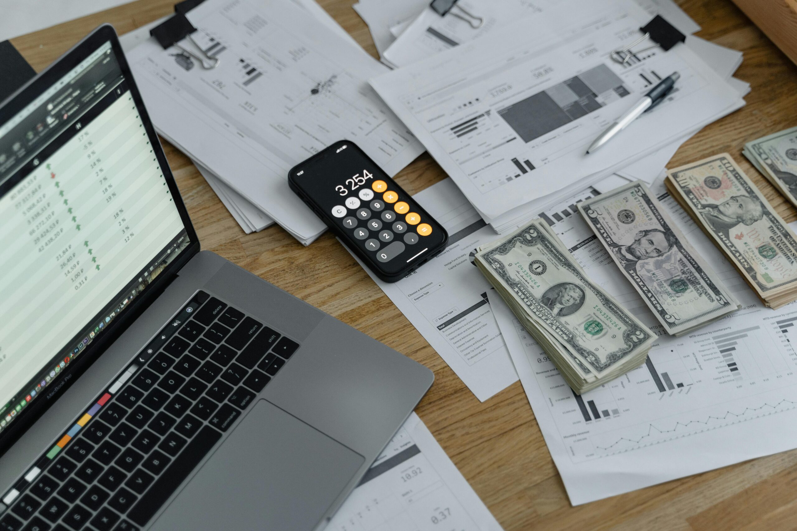 A laptop, smartphone calculator, financial charts, and stacks of U.S. dollar bills on a desk, symbolizing startup costs and financial planning.