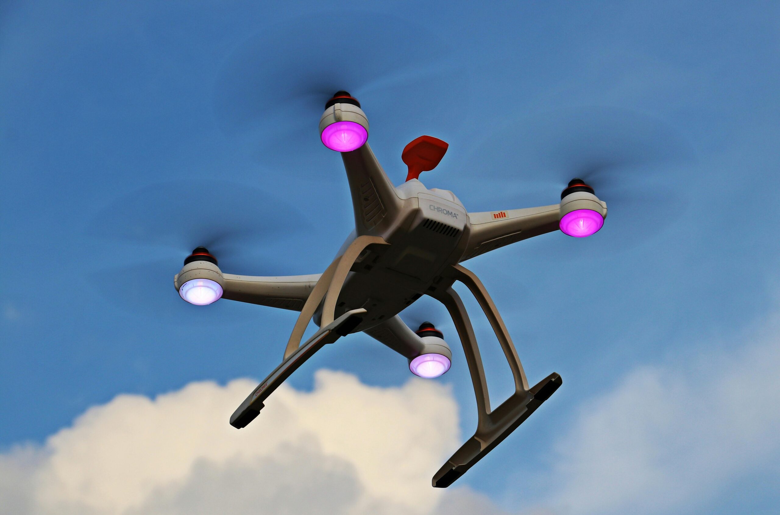 A white quadcopter drone with purple lights flying in the sky above clouds, captured from a low angle.
