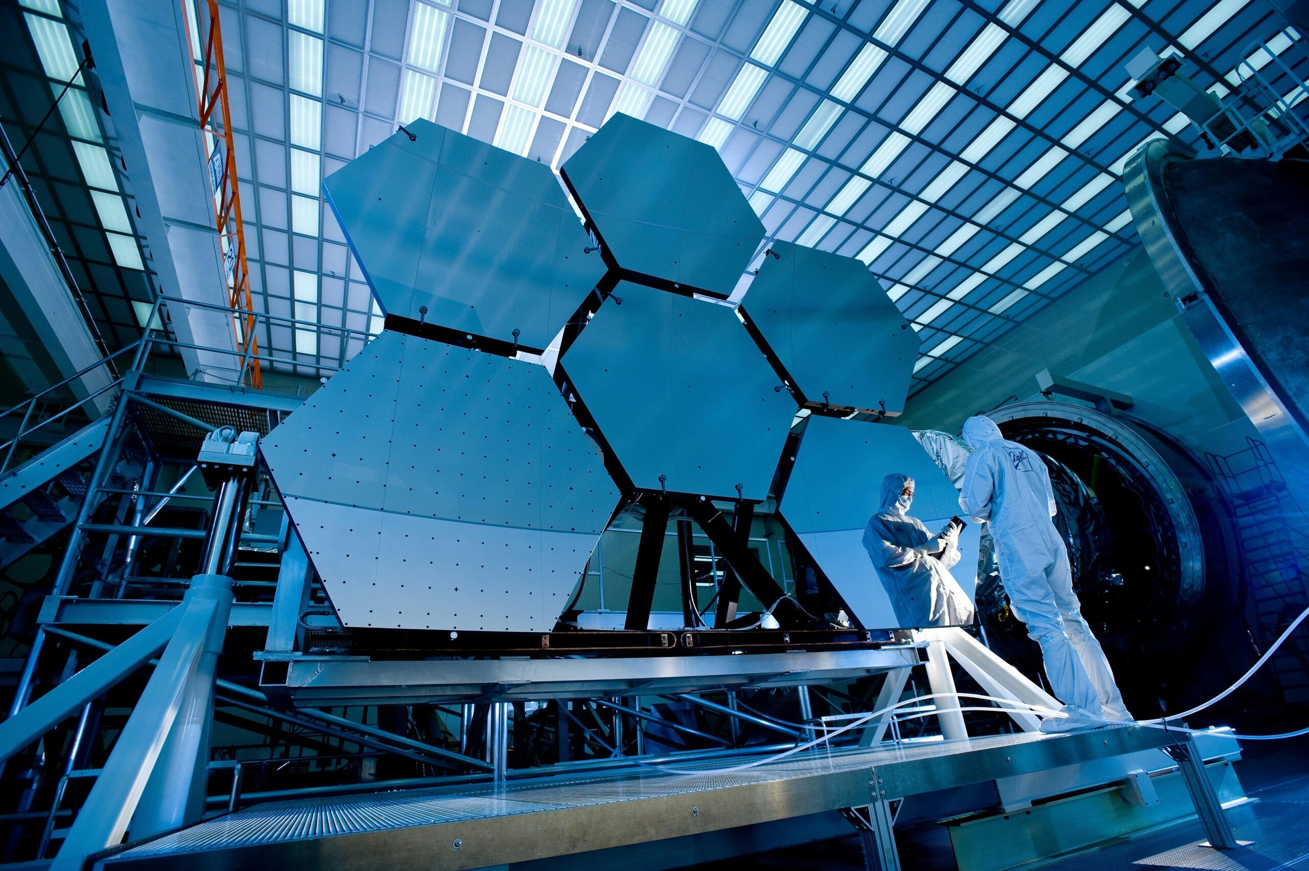 Two scientists in cleanroom suits working on large hexagonal mirror panels inside a high-tech laboratory with bright overhead lighting.